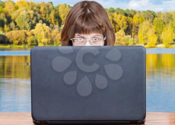 girl with spectacles looks over cover of open laptop with lake on background