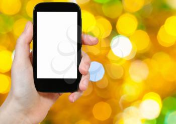 christmas party concept - hand with smartphone with cut out screen on background from yellow and green twinkling Christmas lights bokeh of electric garlands on Xmas tree