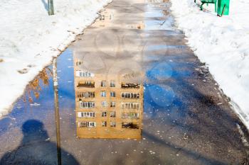 reflection of house in puddle of melting snow on footpath in city in sunny spring day