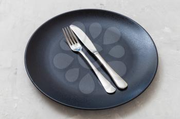 food concept - black plate with parallel knife, spoon on gray concrete surface