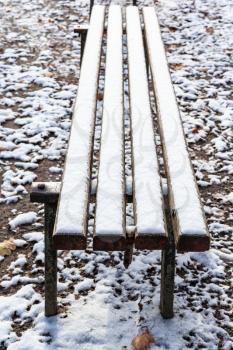 surface of wooden bench covered with the first snow in urban park in frosty autumn day