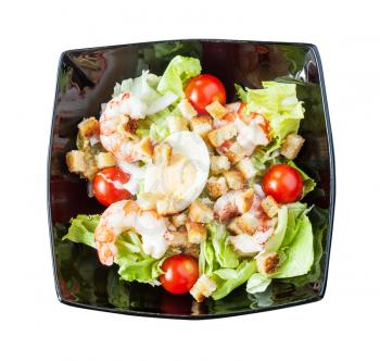 top view of Caesar salad with prawns on black plate isolated on white background