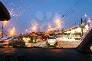 view of cars and street cleaning machines through wet windscreen while driving car on road in Moscow city in winter evening in snow