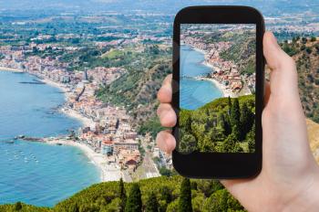 travel concept - tourist photographs Giardini Naxos town of shore of Ionian Sea from Taormina city in Sicily Italy in summer on smartphone