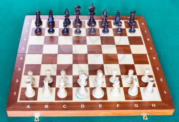 view from white side of wooden chessboard with chess pieces in starting position on green baize table