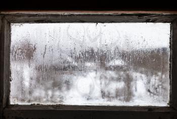 blurry view of russian village through water drops on misted frozen window of rural house in cold winter day (focus on ice surface on the glass)