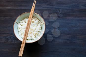 top view of chopsticks above bowl with boiled rice on dark wooden board with copyspace