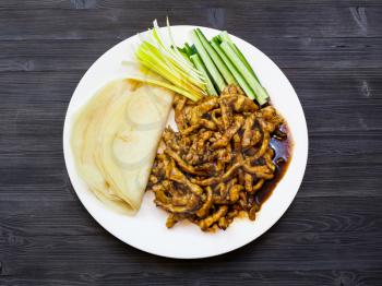 Chinese cuisine dish - top view white plate with sliced pork fried in soy sauce served with spring onion, fresh cucumbers and pancakes (Peking Pork) on dark wooden board