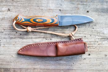 top view of handcrafted blackened steel knife with homemade epoxy and orange fabric handle with hand-sewn leather sheath on old wooden board