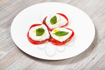 italian cuisine insalata caprese (caprese salad) - stacks from sliced mozzarella cheese, tomato, basil leaf seasoned by olive oil on white plate on wooden table