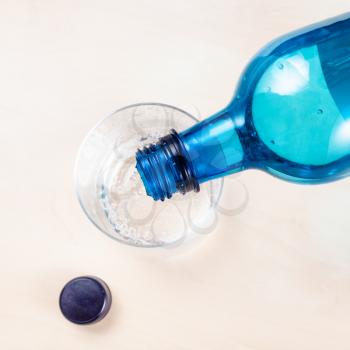 top view of pouring sparkling mineral water from blue plastic bottle in glass on light brown table close up