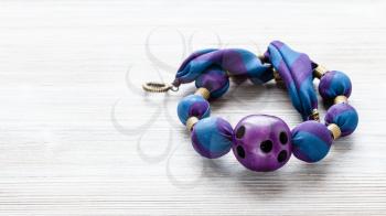 tangled handcrafted necklace of round beads wrapped in blue silk cloth and purple plastic ball with holes on wooden table with copyspace