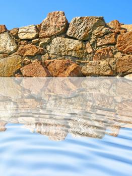 Old stone wall being reflected in ripple blue water.
