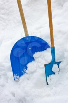 Plastic and metallic blue shovels with wooden handles in the snow heap