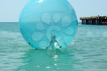 Preschool little girl inside a giant blue bubble-shaped balloon floating on the sea near the pier in a lovely summer day