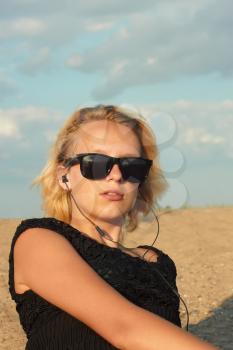 Beautiful teenage blond girl with glasses and headphones on the background of a sandy beach and cloudy sky