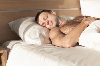 Young European man sleeping in wide bed under white blanket in bright morning sunlight