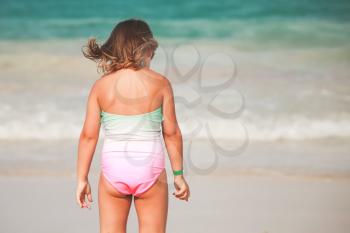 Caucasian blond baby girl in green and pink swimsuit stands on the sea coast, rear view