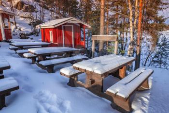 Rural winter landscape with generic red wooden cottage and outdoor furniture under snow, Finland