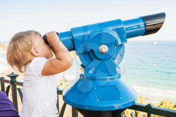 Blond Caucasian baby girl is looking through a stationary paid telescope stands on sea coast 