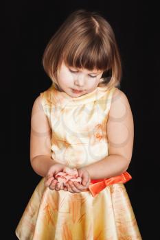 Little blond girl in yellow dress holds pink rose flower made of feathers on black background, vertical studio portrait