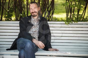 Young bearded happy man on white bench in summer park, outdoor portrait