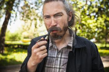 Young serious bearded man smoking pipe in summer park, close-up portrait
