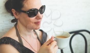 Young adult European woman in dark sunglasses drinks coffee on patio 