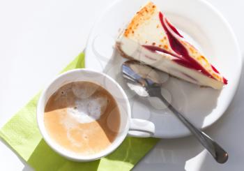 Cup of cappuccino coffee and cheesecake piece with spoon on white saucer. Top view