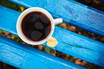 White cup of black tea is on old blue wooden bench in autumn park.  Selective focus with shallow DOF, top view