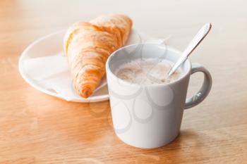 Cappuccino with croissant. Cup of coffee with milk foam stands on wooden table, closeup photo with selective focus