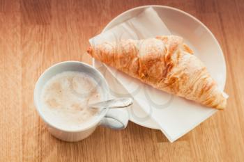 Cappuccino with croissant. Cup of coffee stands on wooden table in cafeteria, closeup photo with selective focus