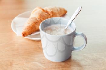 Cappuccino with croissant. Cup of coffee with milk foam stands on wooden table in cafeteria, closeup photo with selective focus