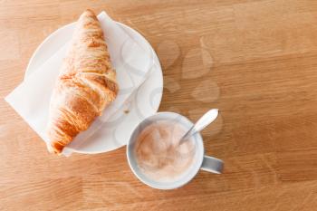 Cappuccino with croissant. Cup of coffee with milk foam stands on wooden table in cafeteria, top view