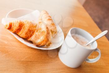 Cappuccino and croissant. Cup of coffee with milk foam stands on wooden table in cafeteria, closeup photo with selective focus