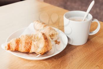 Cappuccino with croissant. Cup of coffee with milk stands on wooden table in cafeteria, closeup photo with selective focus