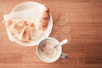 Cappuccino with croissant. Cup of coffee stands on wooden table in cafeteria, closeup photo with selective focus, top view, warm tonal correction filter effect