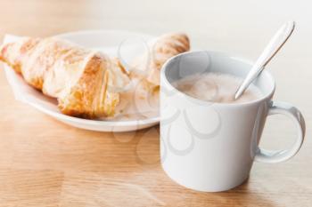 Breakfast menu. Cappuccino with croissant. Cup of coffee with milk foam stands on wooden table in cafeteria, closeup photo with selective focus
