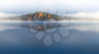 Autumnal nature panoramic landscape. Island on still lake in cold foggy morning