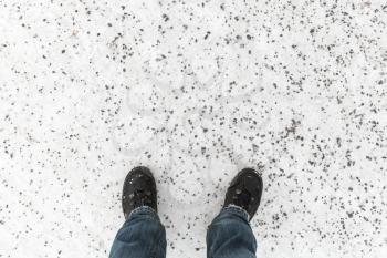Male feet in blue jeans and black shoes standing on snowy winter road with anti-slip granite chippings, first person view 