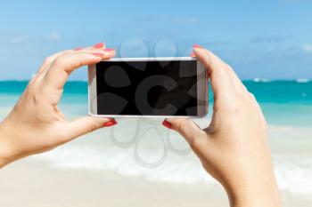 Woman holds smart phone in hands for taking outdoor photo on a summer beach in Dominican republic