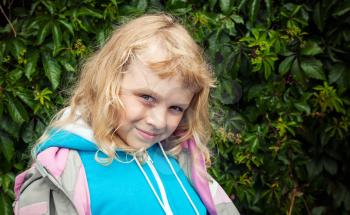 Little blond girl wears casual sport clothes. Outdoor smiling portrait above green leaves background