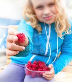 Little blond active girl held out fresh raspberry from his full box. Outdoor portrait with selective focus on berry