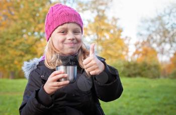 little beautiful blond girl drinks tea in autumnal park and showing thumbs up