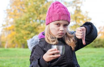 little beautiful blond girl drinks tea in autumnal park and showing thumbs down