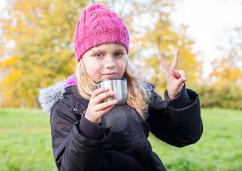little beautiful blond girl drinks tea in autumnal park and showing victory fingers