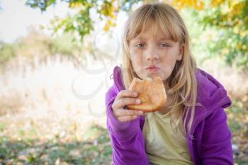 Little blond girl in autumn park eats small pie
