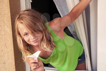 Little blond Caucasian girl with paper plane in the window, outdoor closeup portrait