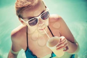 Little blond girl drinks cocktail in a swimming pool, vintage toned photo filter effect