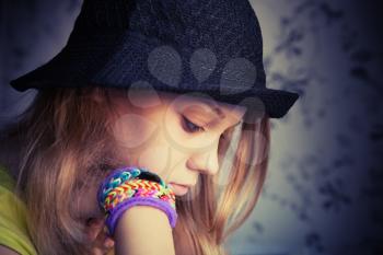 Profile portrait of beautiful blond teenage girl in black hat and rubber loom bracelets, vintage dark toned photo, instagram style effect